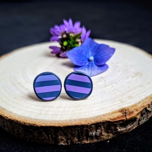 Op de afbeelding: Twee ronde oorstekers met een donkerblauwe basis en horizontale paarse strepen. De oorbellen worden gepresenteerd op een houten schijf met paarse en blauwe bloemen op de achtergrond. Sieraden.