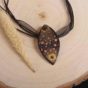 Op de afbeelding: Een zwarte en gouden ketting met een geometrisch ontwerp. De hanger is druppelvormig en heeft een zwarte achtergrond met gouden accenten. De ketting is gemaakt van zwart lint.