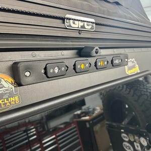 May include: Black vehicle roof with a row of LED lights. The lights are mounted on a black bar above the window. A sticker with the text "STATICLINE OVERLAND" is visible. An American flag is in the background.