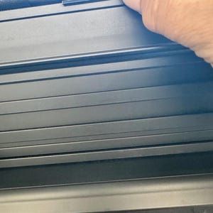May include: Close-up of a black, textured, multi-slat surface. A hand is touching the top edge of the surface. The surface appears to be part of a larger object, possibly a cover or a panel.