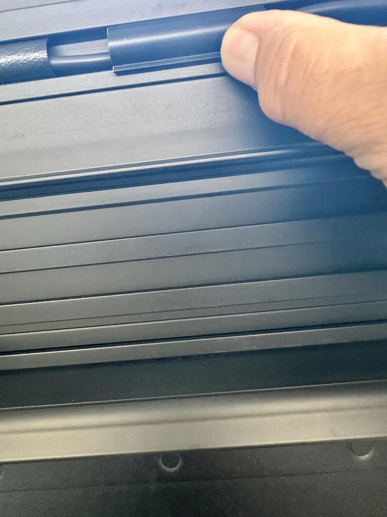 May include: Close-up of a black, textured, multi-slat surface. A hand is touching the top edge of the surface. The surface appears to be part of a larger object, possibly a cover or a panel.