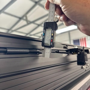 May include: A digital caliper measuring the thickness of a black, grooved surface. The caliper reads 0.4815 inches. The image shows a close-up of the measurement process, highlighting the precision of the tool.
