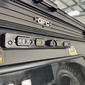 May include: Black vehicle roof rack with a row of small LED lights. The rack has a GFC logo and a sticker with a mountain and vehicle graphic. The lights are in black housings with clear and amber lenses.