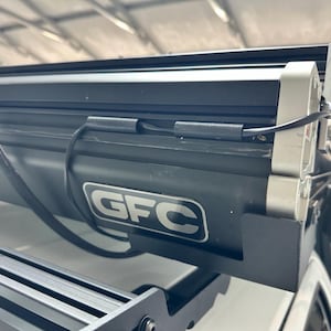 May include: A black GFC roof rack with a silver accent and the GFC logo. The rack is mounted on a white vehicle and includes a black light bar and wiring.