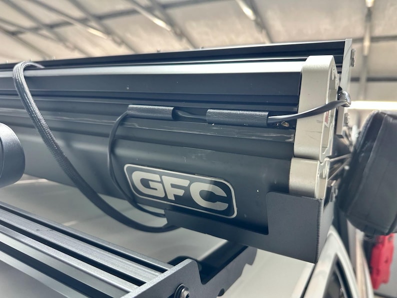 May include: A black GFC roof rack with a silver accent and the GFC logo. The rack is mounted on a white vehicle and includes a black light bar and wiring.