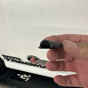 May include: A section of black rubber trim, likely for a vehicle, held in a hand. The trim has a rounded top and a flat base. The background shows a white vehicle panel with the Toyota Tacoma logo and a 4 Wheel Drive emblem.