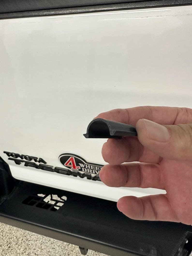 May include: A section of black rubber trim, likely for a vehicle, held in a hand. The trim has a rounded top and a flat base. The background shows a white vehicle panel with the Toyota Tacoma logo and a 4 Wheel Drive emblem.