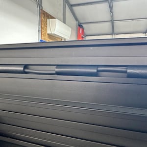 May include: Close-up of a black, metallic, roll-up truck bed cover. The cover is composed of multiple horizontal slats with a black rubber tube running through the center. The background includes a garage setting.