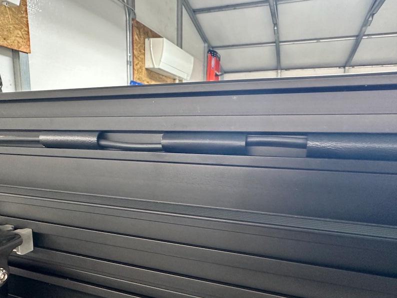 May include: Close-up of a black, metallic, roll-up truck bed cover. The cover is composed of multiple horizontal slats with a black rubber tube running through the center. The background includes a garage setting.
