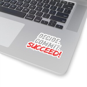 May include: A white sticker with the words "DECIDE. COMMIT. SUCCEED!" in gray and red lettering. The sticker is on a silver laptop, near the keyboard.
