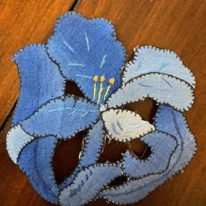May include: A blue fabric flower with a yellow center, made using the patchwork technique. The flower is stitched together with black thread.