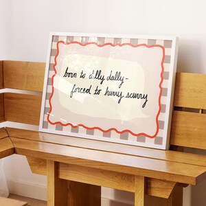 K&ouml;nnte beinhalten: Ein wei&szlig;er gerahmter Druck mit einem rot-wei&szlig; karierten Hintergrund. Der Druck zeigt eine wei&szlig;e Sprechblase mit dem Text "born to dally dally - forced to hurry scurry".