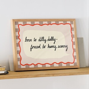 K&ouml;nnte beinhalten: Ein gerahmter Druck mit wei&szlig;em Hintergrund und rotem und schwarzem kariertem Rand. Der Druck zeigt ein handgeschriebenes Zitat, das lautet "born to dilly dally - forced to hurry scurry".