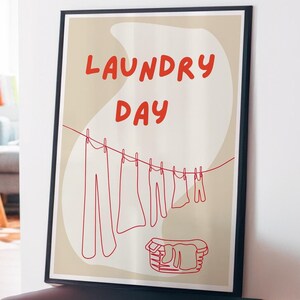 May include: A beige poster with the text "Laundry Day" in red. A line of red clothes hang on a clothesline, including pants, shirts, and a few smaller items. A red basket sits below the clothesline.