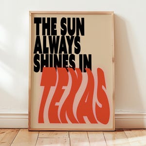 May include: A framed poster with the text "THE SUN ALWAYS SHINES IN TEXAS" in black and orange. The poster has a beige background and a wooden frame, leaning against a white wall and wooden floor.