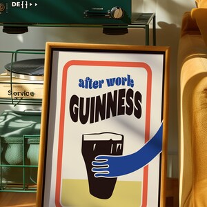 Op de afbeelding: Een ingelijste poster met een witte achtergrond en rode, oranje en gele randen. De poster toont een zwart-witte illustratie van een pint Guinness met een blauwe hand die ernaar reikt. De tekst "after work GUINNESS" is in zwart-wit gedrukt.
