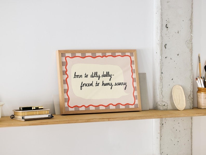 K&ouml;nnte beinhalten: Ein gerahmter Druck mit wei&szlig;em Hintergrund und rotem und schwarzem kariertem Rand. Der Druck zeigt ein handgeschriebenes Zitat, das lautet "born to dilly dally - forced to hurry scurry".