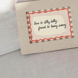 K&ouml;nnte beinhalten: Ein beigefarbenes Schild mit einem roten und wei&szlig;en karierten Rahmen. Das Schild hat eine wei&szlig;e Sprechblase mit dem Text "born to dilly dally - forced to hurry scurry" in schwarzer Schrift.