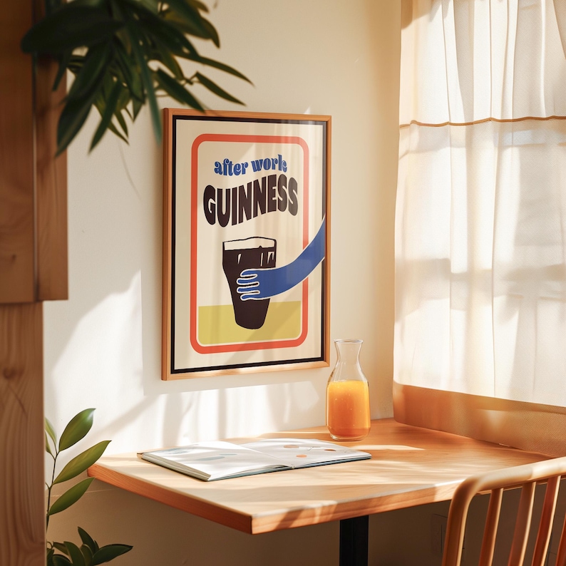 Op de afbeelding: Een ingelijste poster met een vintage ontwerp met een glas Guinness-bier en de tekst "after work GUINNESS". De poster hangt aan een witte muur boven een houten tafel met een glas sinaasappelsap.