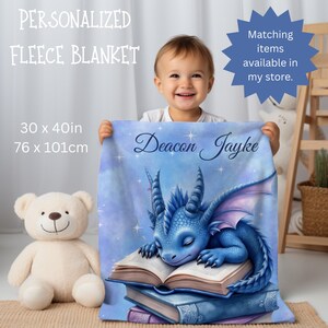 May include: A personalised fleece blanket, 76 x 101cm, displays a sleeping blue dragon on books. The blanket is inscribed with the name "Deacon Jayke". The background is a soft blue with stars.