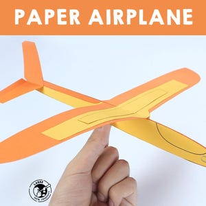 Może przedstawiać: Pomarańczowo-żółty papierowy samolot jest zrównoważony na palcu. Rozpiętość skrzydeł samolotu wynosi około 25 cm. Słowa "PAPER AIRPLANE" są dużymi białymi literami u góry obrazu.