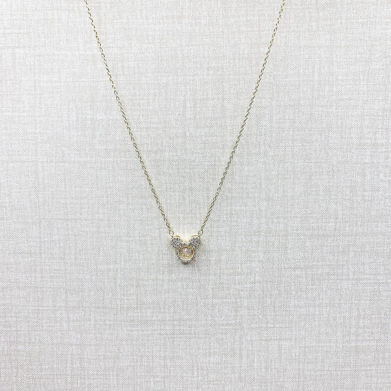 K&ouml;nnte beinhalten: Eine goldfarbene Halskette mit einem Anh&auml;nger in Form eines Cartoon-Mauskopfes. Der Anh&auml;nger ist mit kleinen, klaren Kristallen besetzt. Die zarte Kette h&auml;ngt vor einem neutralen Hintergrund.