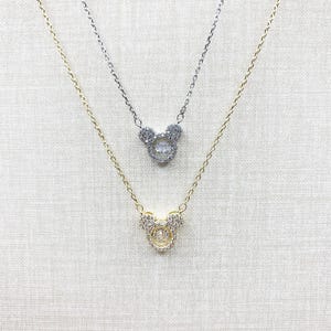 K&ouml;nnte beinhalten: Zwei Halsketten mit Anh&auml;ngern in Form eines Mickey Mouse-Kopfes. Eine Halskette ist goldfarben, die andere silberfarben. Die Anh&auml;nger sind mit kleinen, klaren Kristallen besetzt. Die zarten Ketten h&auml;ngen vor einem neutralen Hintergrund.