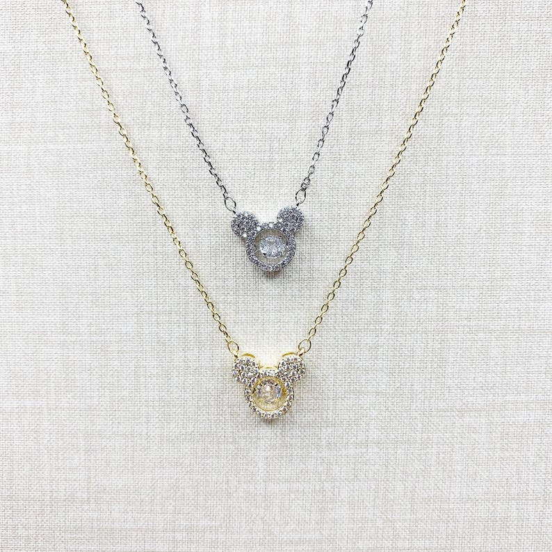 K&ouml;nnte beinhalten: Zwei Halsketten mit Anh&auml;ngern in Form eines Mickey Mouse-Kopfes. Eine Halskette ist goldfarben, die andere silberfarben. Die Anh&auml;nger sind mit kleinen, klaren Kristallen besetzt. Die zarten Ketten h&auml;ngen vor einem neutralen Hintergrund.