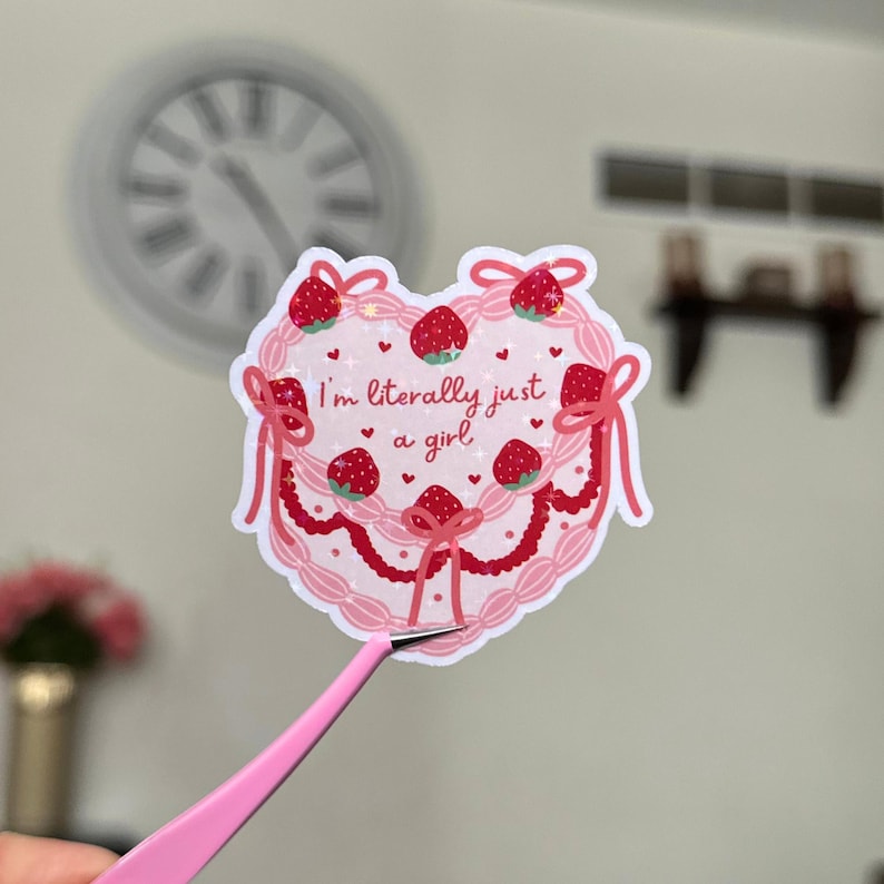 Puede incluir: Pegatina rosa en forma de coraz&oacute;n con un dise&ntilde;o de pastel rosa y blanco, decorado con fresas rojas y lazos rosas. El texto "I'm literally just a girl" est&aacute; escrito en negro en la pegatina.