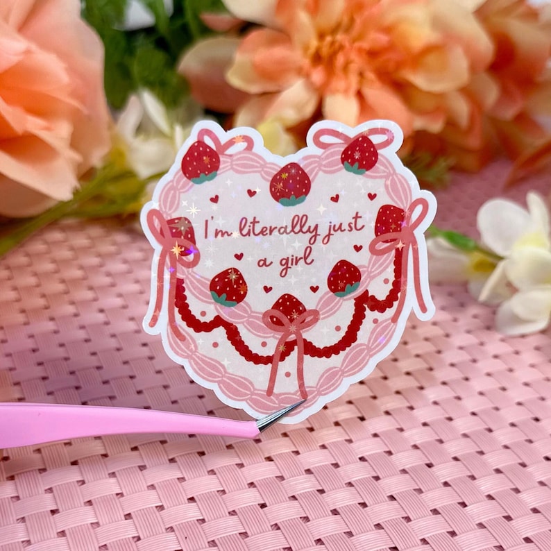 Puede incluir: Una pegatina rosa en forma de coraz&oacute;n con un dise&ntilde;o de pastel blanco y fresas rojas. La pegatina tiene brillo y el texto "I'm literally just a girl" en negro.