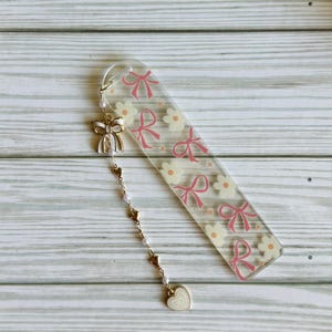 May include: Clear acrylic bookmark with a pink bow and flower pattern. A gold-colored charm with a white bow, hearts, pearls, and a heart-shaped pendant hangs from the top. The bookmark is approximately 6 inches long.
