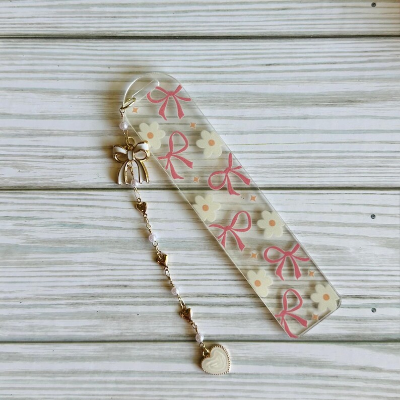 May include: Clear acrylic bookmark with a pink bow and flower pattern. A gold-colored charm with a white bow, hearts, pearls, and a heart-shaped pendant hangs from the top. The bookmark is approximately 6 inches long.