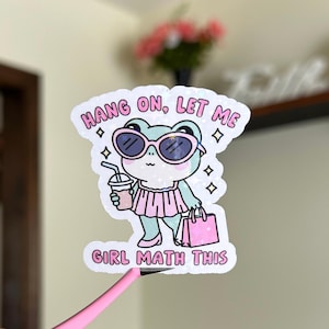 May include: A sticker featuring a cartoon frog wearing sunglasses and a pink dress, holding a pink shopping bag and a cup of coffee. The text on the sticker reads "Hang on, let me girl math this."