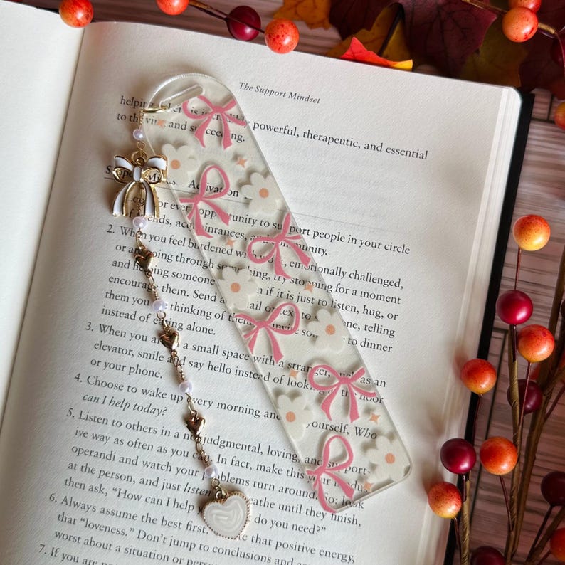 May include: Clear acrylic bookmark with pink bow and daisy pattern. A gold-colored charm with a white bow and pearl accents hangs from the top. A heart-shaped charm dangles from the bottom. The bookmark is placed on an open book.