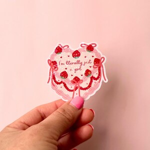 Puede incluir: Una pegatina rosa en forma de coraz&oacute;n con un dise&ntilde;o de pastel rosa y blanco. El pastel est&aacute; decorado con fresas y el texto "I'm literally just a girl".