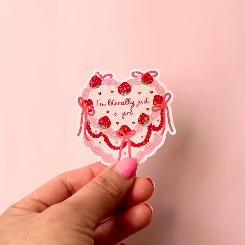 Puede incluir: Una pegatina rosa en forma de coraz&oacute;n con un dise&ntilde;o de pastel rosa y blanco. El pastel est&aacute; decorado con fresas y el texto "I'm literally just a girl".