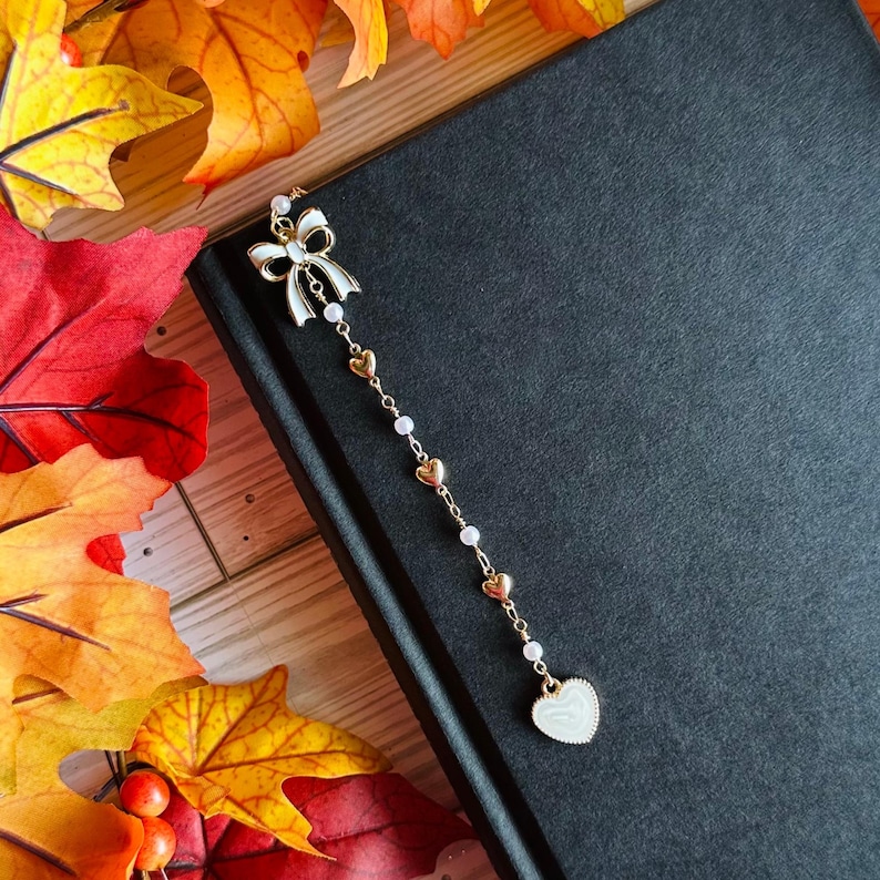 May include: A gold-colored bookmark with a white bow and a chain of small gold hearts and pearls. The bookmark is resting on a closed black book. A heart-shaped charm hangs at the end of the chain. Autumn leaves in red, orange, and yellow surround the book.