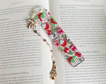 May include: Clear acrylic bookmark with a Christmas theme. The bookmark features festive illustrations of wrapped gifts, hot cocoa, books, and bows in red, green, and white. A charm with a Christmas tree and pearls hangs from the bottom.