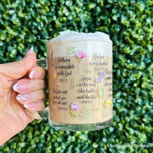 May include: A clear glass coffee mug filled with a creamy beverage and topped with whipped cream. The mug is decorated with floral designs and inspirational quotes from the Bible. The mug is held by a person.
