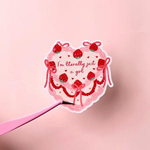Puede incluir: Pegatina rosa en forma de coraz&oacute;n con un dise&ntilde;o de pastel rosa y blanco. El pastel est&aacute; decorado con fresas rojas y lazos blancos. El texto "I'm literally just a girl" est&aacute; escrito en rosa en el pastel.