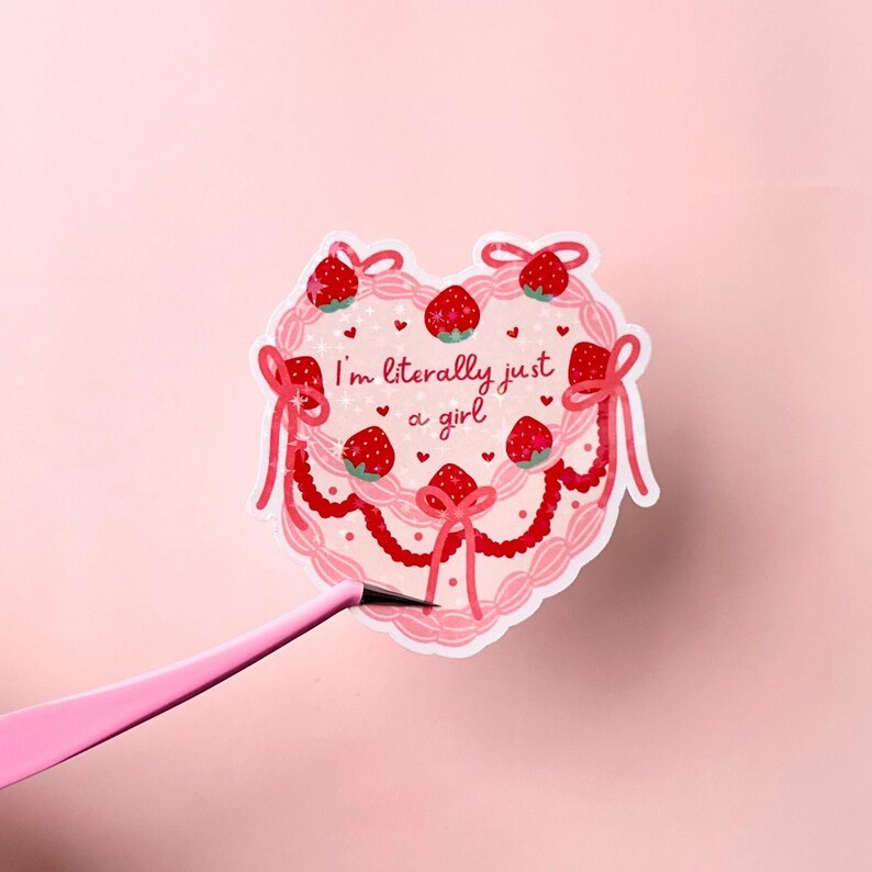 Puede incluir: Pegatina rosa en forma de coraz&oacute;n con un dise&ntilde;o de pastel rosa y blanco. El pastel est&aacute; decorado con fresas rojas y lazos blancos. El texto "I'm literally just a girl" est&aacute; escrito en rosa en el pastel.