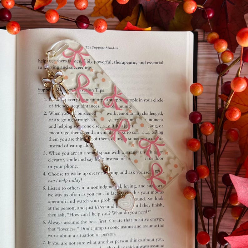 May include: Clear acrylic bookmark with a floral and bow design. The bookmark has a gold chain with a heart charm. The bookmark is resting on an open book with text.
