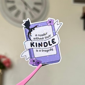 May include: A sticker featuring a purple book design with a dragon silhouette and the text "a reader without their KINDLE is a tragedy." The sticker also includes purple flower accents and a white banner.