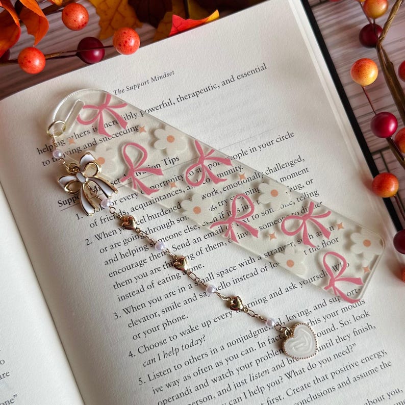 May include: A clear acrylic bookmark with pink bow and daisy patterns. The bookmark has a gold chain with heart and pearl charms. The text "The Support Mindset" is visible on the page.