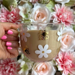 May include: A glass mug with a white daisy and bee design. The mug is filled with a light brown beverage.
