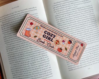 May include: A bookmark for the Cozy Girl Book Club, featuring fall-themed illustrations like boots, an umbrella, and a pumpkin. The bookmark is on a book with text.