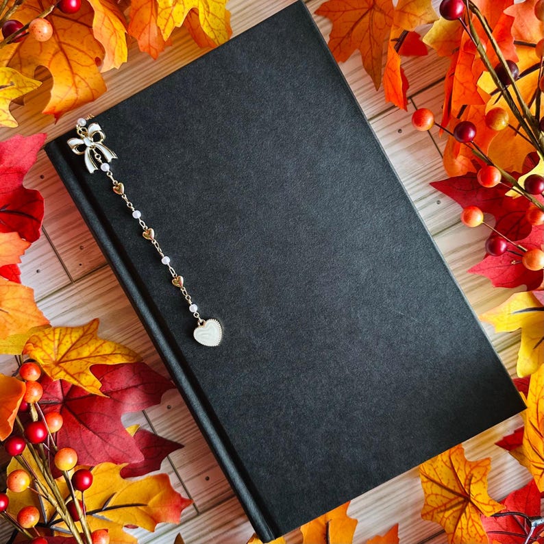 May include: A black book with a decorative bookmark featuring a white bow, gold hearts, and a heart-shaped charm. The book is surrounded by colorful autumn leaves and berries, creating a seasonal theme.