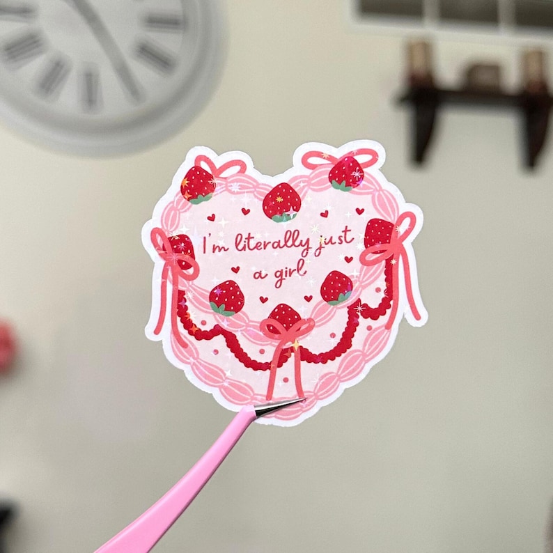 Puede incluir: Pegatina rosa en forma de coraz&oacute;n con un dise&ntilde;o de pastel rosa y blanco. El pastel est&aacute; decorado con fresas y lazos rosas. El texto "I'm literally just a girl" est&aacute; escrito en el pastel.