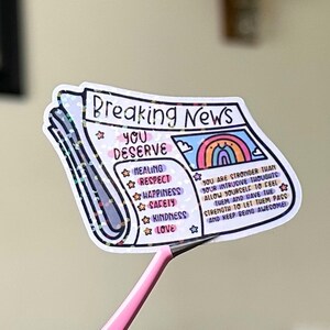May include: A sticker with a newspaper design that says "Breaking News: You Deserve" with a rainbow and a list of positive affirmations including healing, respect, happiness, safety, kindness, and love. The sticker has a glittery finish.