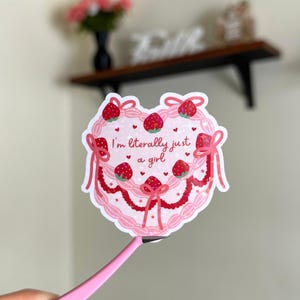 Puede incluir: Pegatina rosa en forma de coraz&oacute;n con un dise&ntilde;o de pastel rosa y blanco. El pastel est&aacute; decorado con fresas y el texto "Soy literalmente solo una chica".